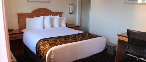 A hotel room with a large bed, a desk, and a chair. There is a door leading to another room and a trash can.