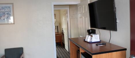 A hotel room with a wooden desk, a television mounted on the wall, a chair, and a door leading to a bathroom.