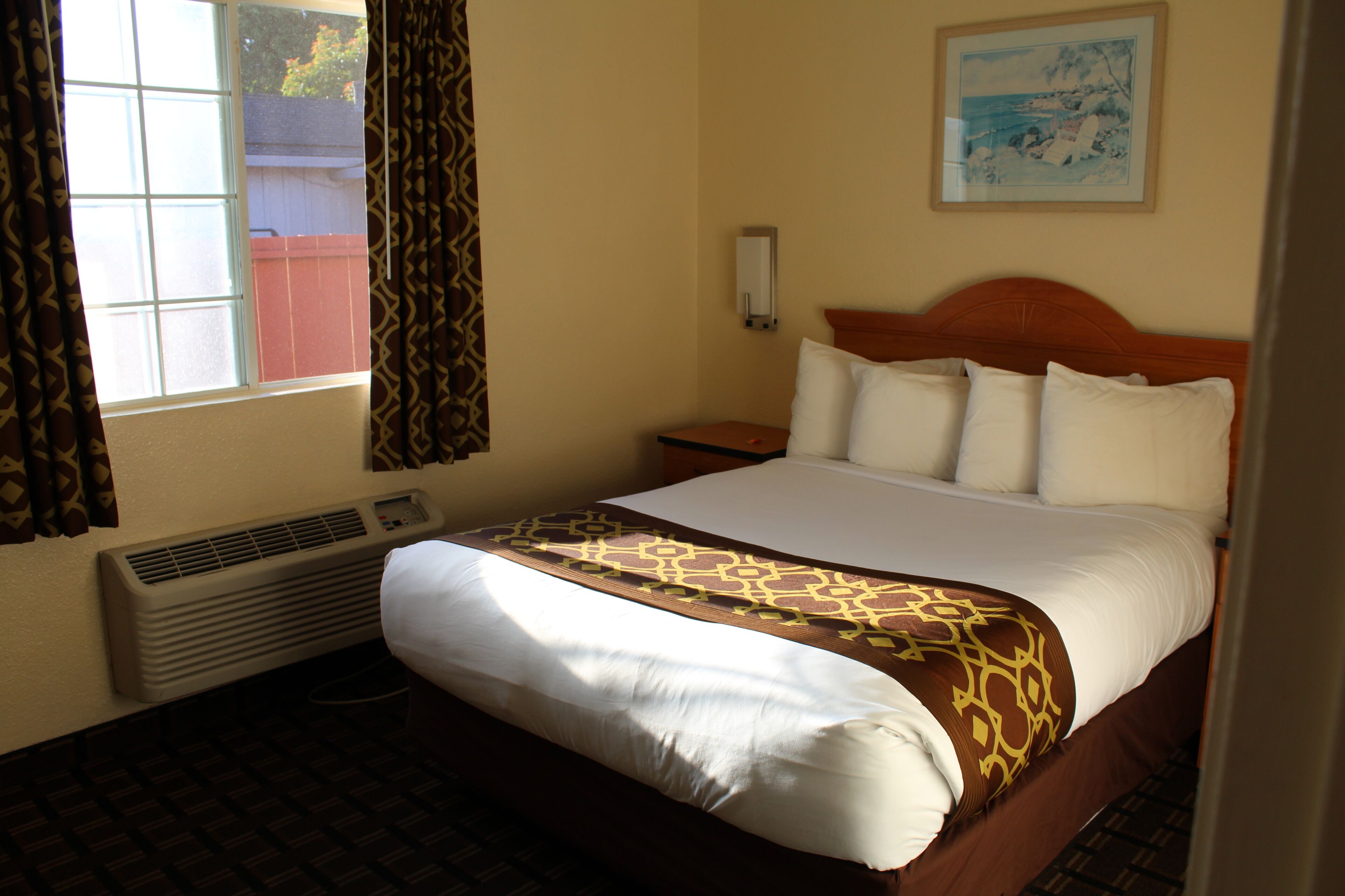 A hotel room with a bed, a nightstand, a wall-mounted lamp, a window with curtains, and a painting on the wall.
