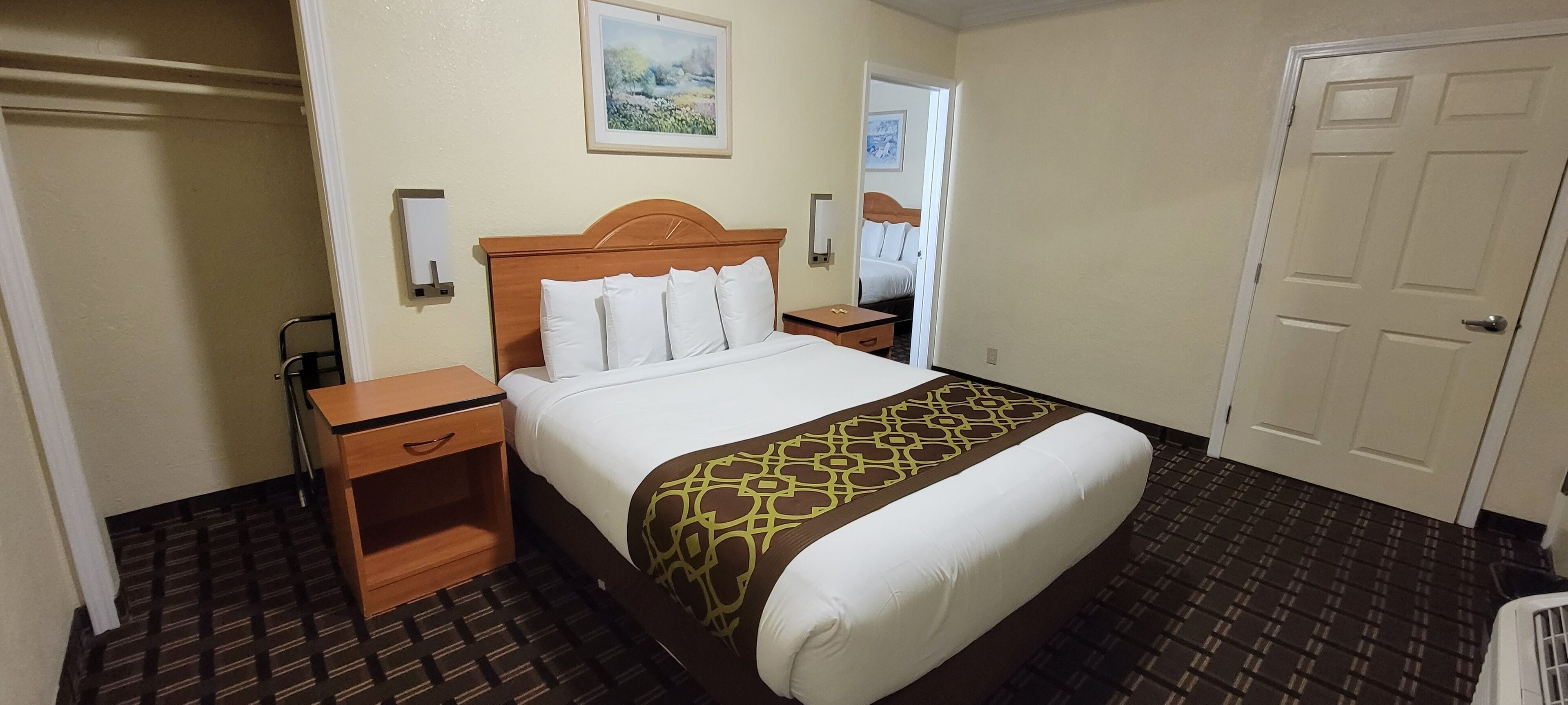 Une chambre d’hôtel avec un lit, une table de chevet en bois, une petite table, une lampe fixée au mur et une porte.