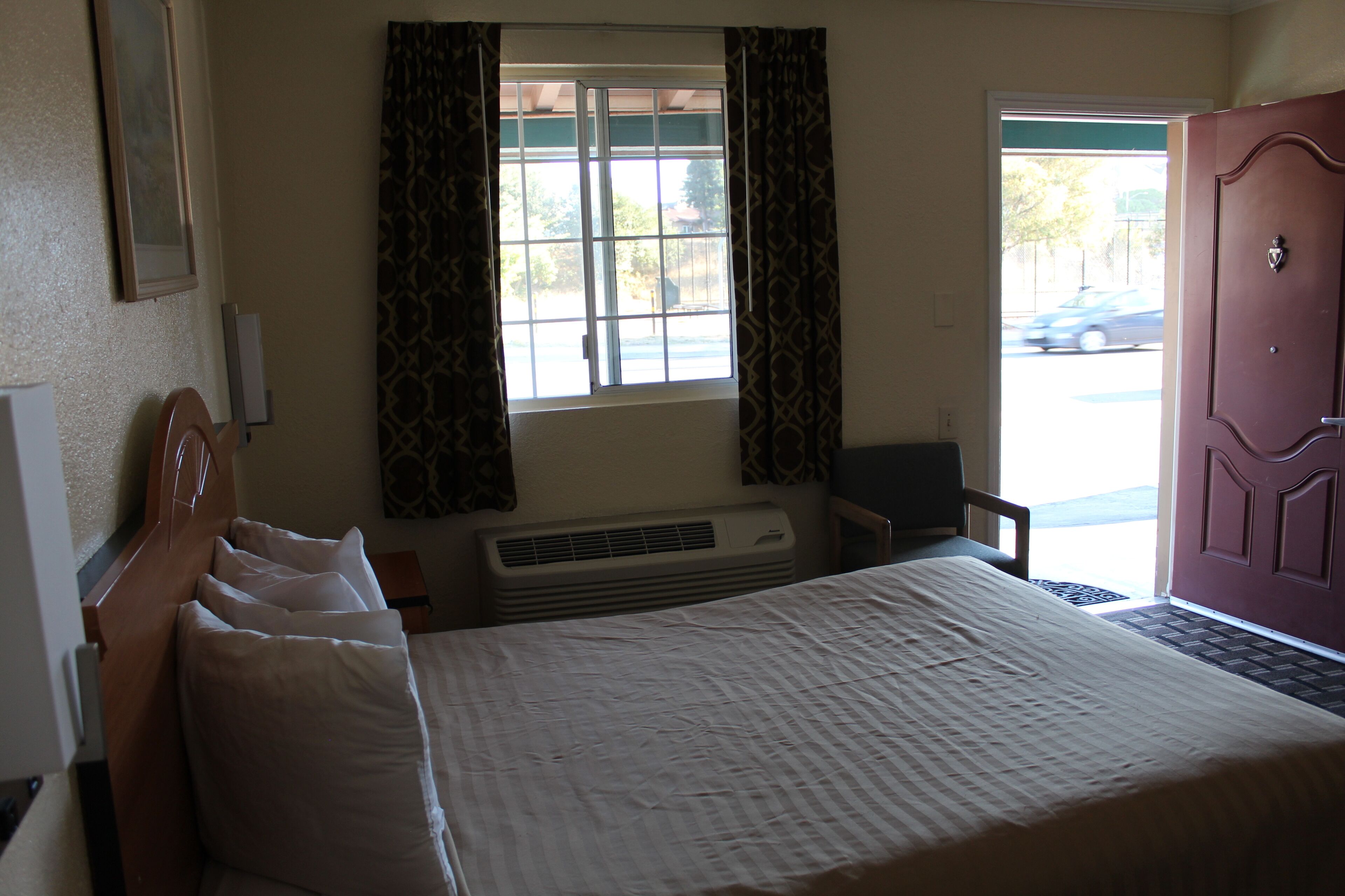 Een hotelkamer met een bed, een raam met gordijnen, een airconditioning en een deur, plus uitzicht op de straat.