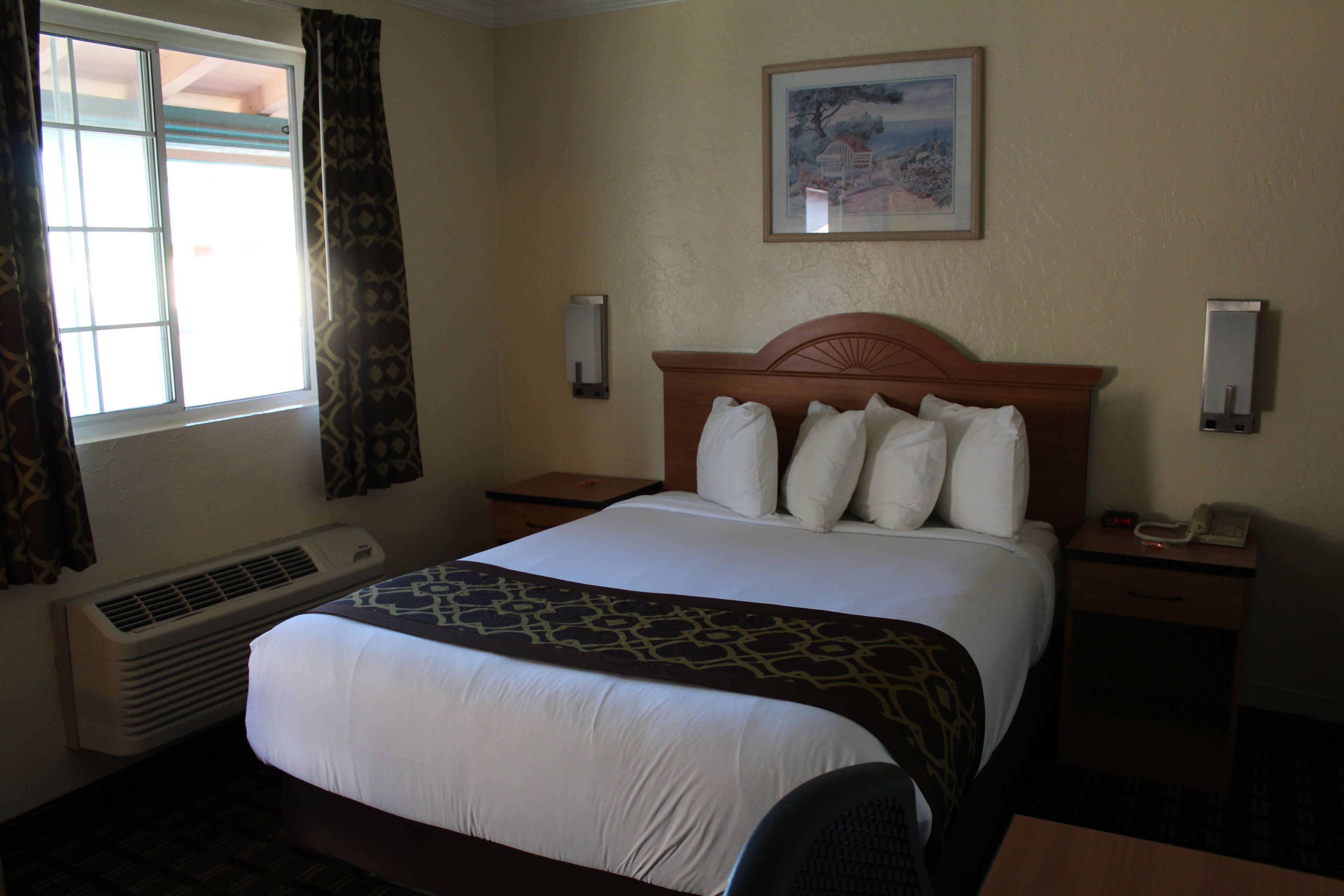 A hotel room with a large bed, a bedside table with a phone, a wall-mounted air conditioner, and a painting above the bed.