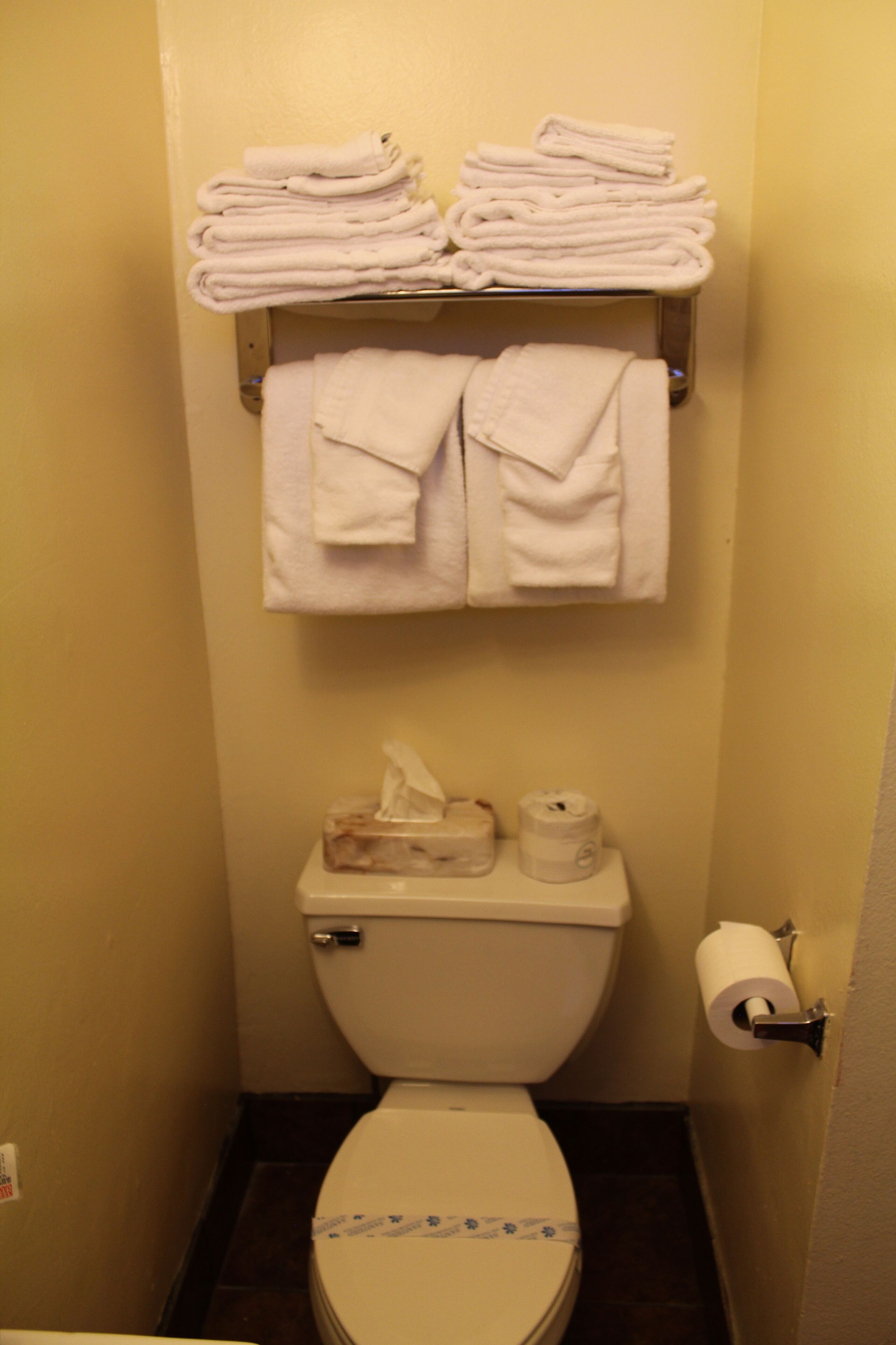 A small bathroom with a toilet, a stack of white towels, a tissue box, and a toilet paper holder.