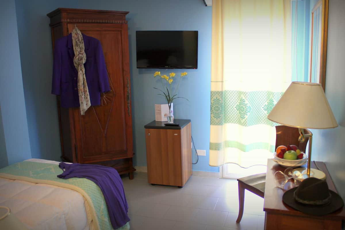Habitación de hotel con cama, armario, pequeño mueble de madera, televisor, ventana con cortinas, mesa con lámpara y un frutero con fruta.