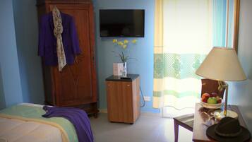 Habitación de hotel con cama, armario, pequeño mueble de madera, televisor, ventana con cortinas, mesa con lámpara y un frutero con fruta.
