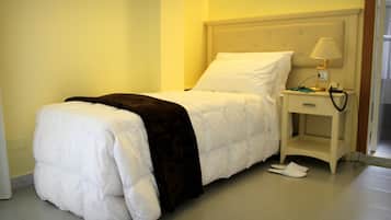 Una cama bien hecha con cubrecama blanco y manta oscura, una mesita de noche con lámpara y teléfono, y un par de zapatos en el suelo.