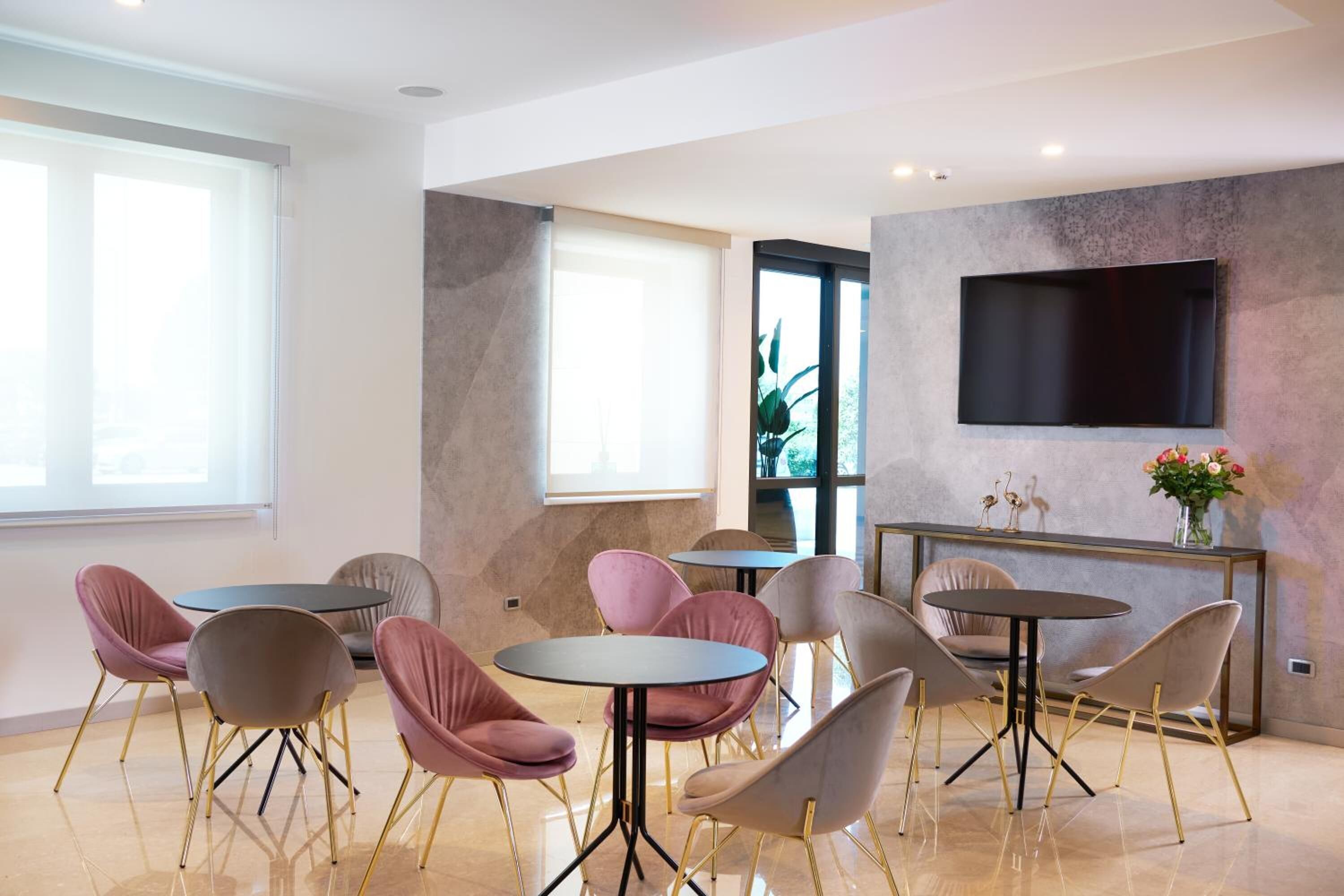 A modern lounge area with round tables and chairs, a flat-screen TV mounted on the wall, and a large window providing natural light.