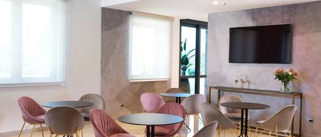 A modern lounge area with round tables and chairs, a flat-screen TV mounted on the wall, and a large window providing natural light.
