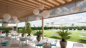 Un área de comedor al aire libre con mesas y sillas redondas, plantas en macetas y sombrillas.