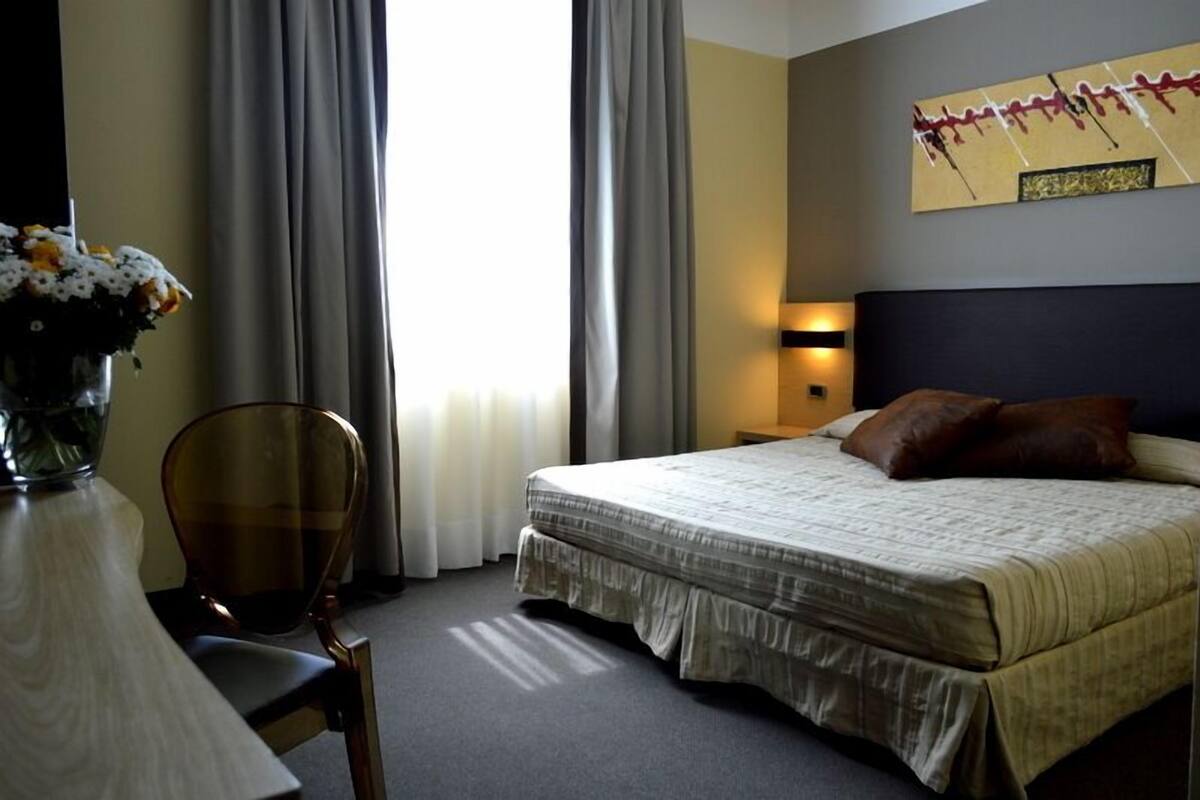 Una habitación de hotel con una cama, una silla, un escritorio, un florero y un cuadro en la pared.