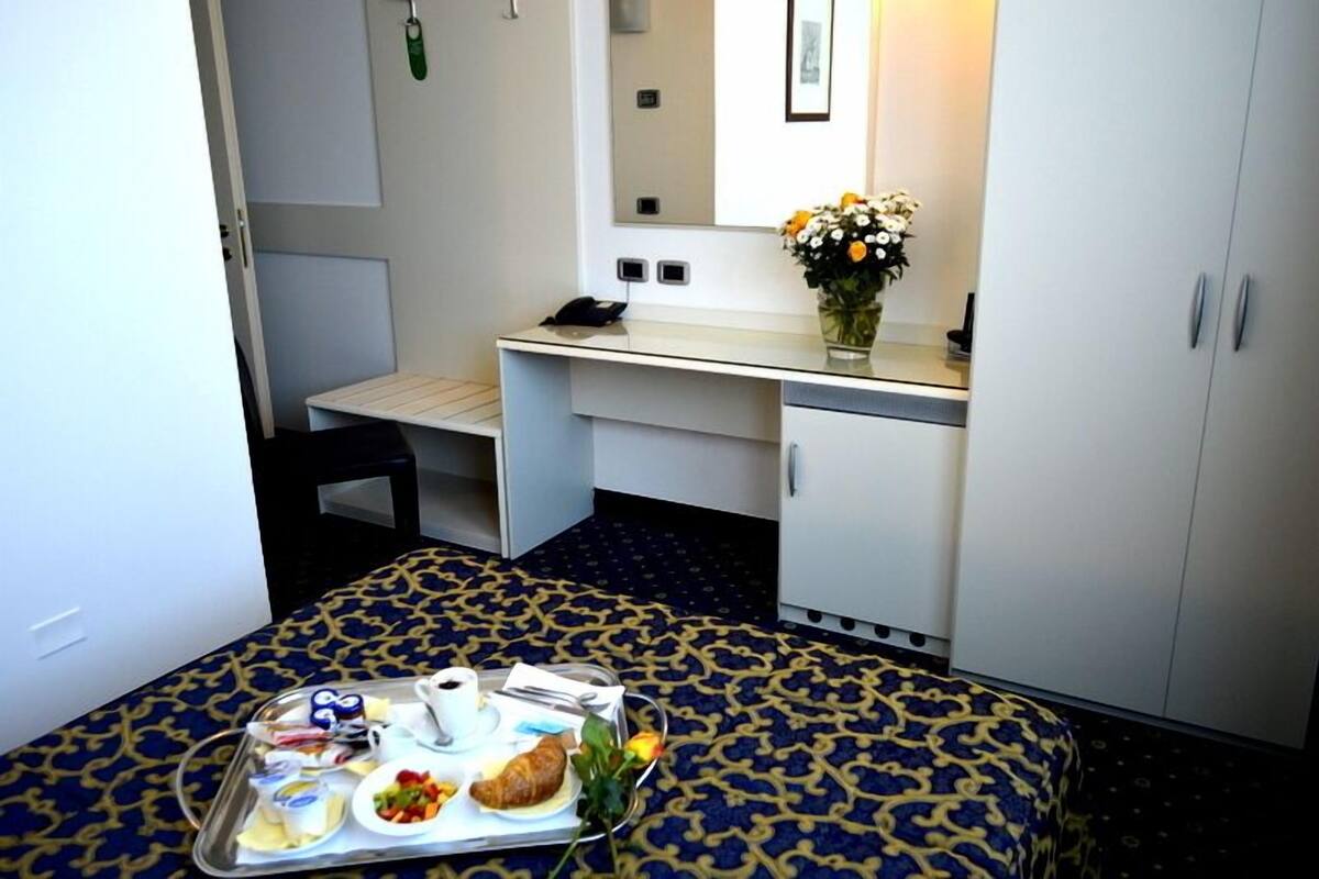 Habitación de hotel pequeña con cama, escritorio, silla, mesita con alimentos para el desayuno, espejo y armario.