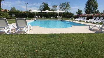 Ein Schwimmbecken mit weißen Liegestühlen und Sonnenschirmen, umgeben von Gras und Bäumen.