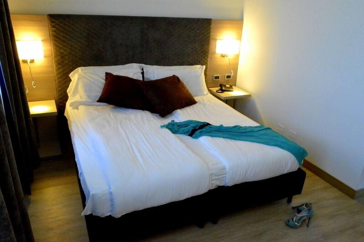 Una cama bien hecha con sábanas blancas y una manta color turquesa, dos almohadas oscuras y un cabecero de madera. Hay dos lámparas de noche a cada lado de la cama, y se ven un par de zapatos en el suelo.