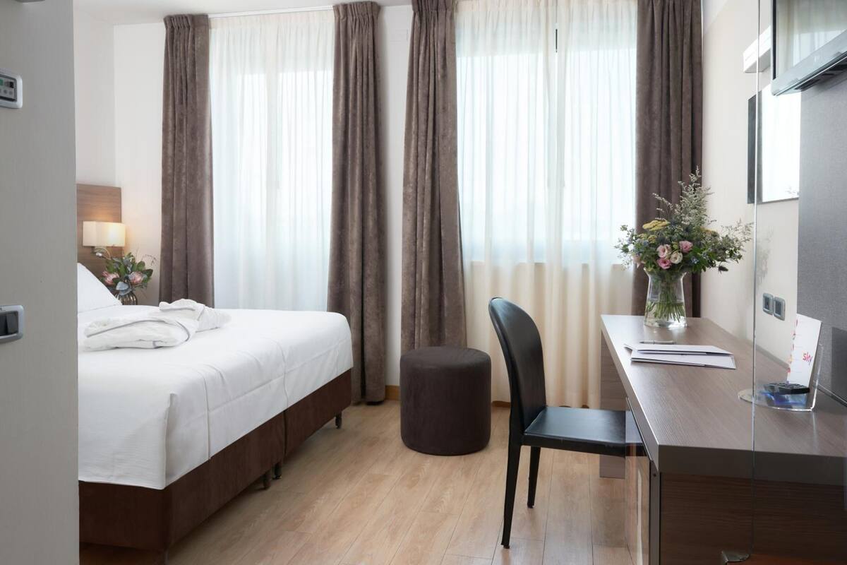 Habitación de hotel con cama, escritorio y silla, un pequeño reposapiés y un florero con flores.