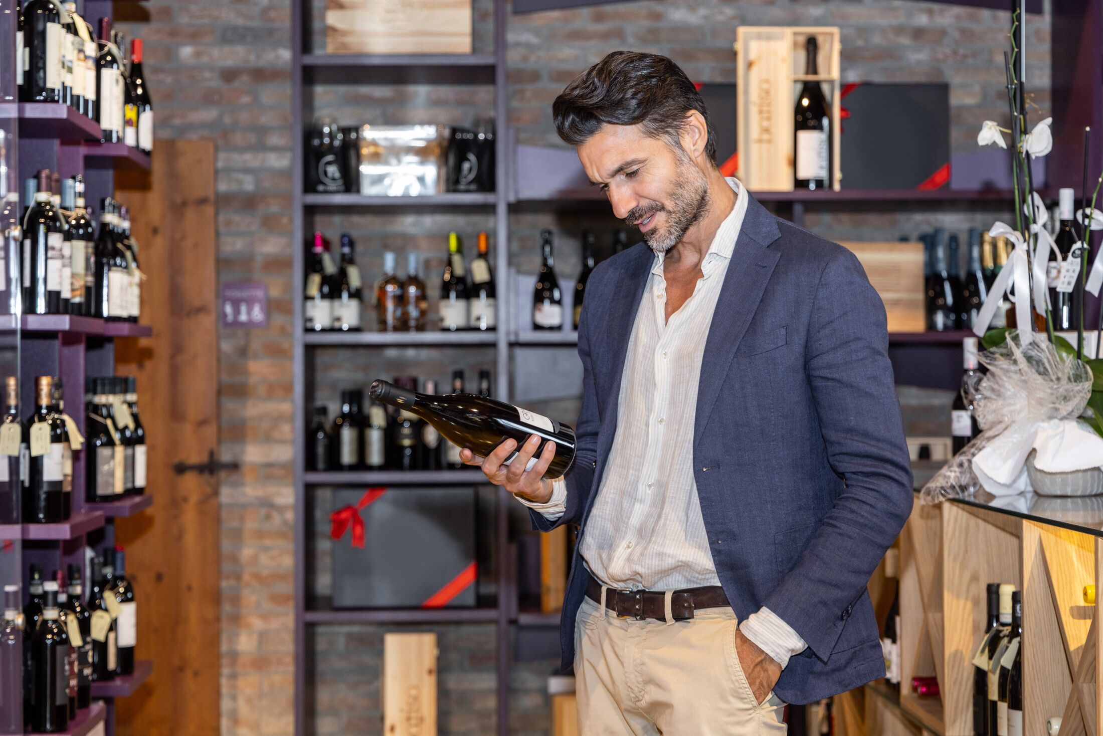A man holding a wine bottle in a wine shop with shelves of various bottles and a wooden crate in the background.