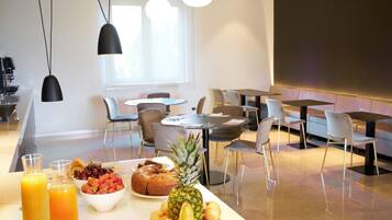 Um buffet de café da manhã com frutas, doces e sucos em uma área de refeições moderna de hotel, com móveis contemporâneos e iluminação pendente.