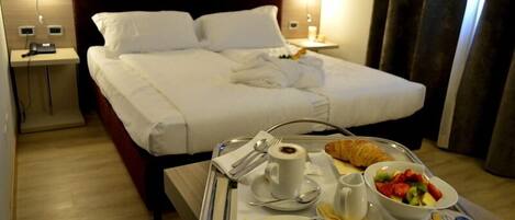 A hotel room with a bed, bedside tables, and a breakfast tray with croissants, fruit, and coffee.
