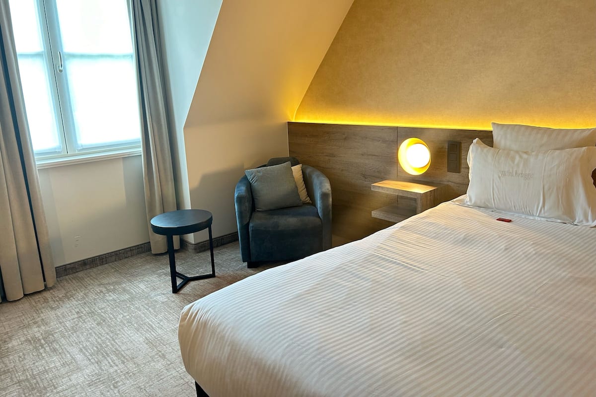 Habitación de hotel con una cama grande, una mesita redonda pequeña, una silla, una lámpara de noche y un ventanal con cortinas.