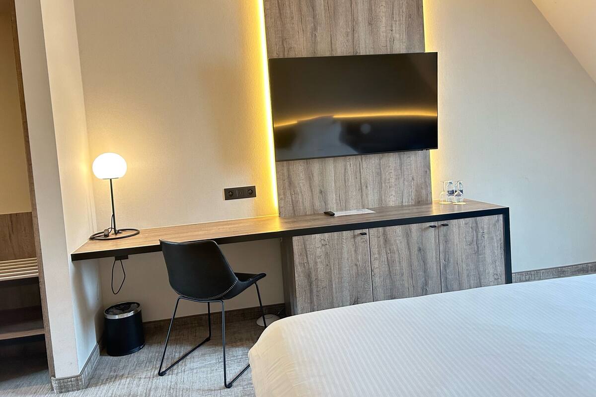 Una habitación de hotel moderna con un televisor de pantalla plana montado en una pared de paneles de madera, un escritorio con una lámpara, una silla negra y una cama.