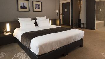 Ein modernes Hotelzimmer mit einem großen Bett, einem dunklen Kopfteil und Teppichboden. An den Wänden hängen zwei gerahmte Bilder, ein Nachttisch mit Lampe und ein kleiner Tisch mit Stuhl.