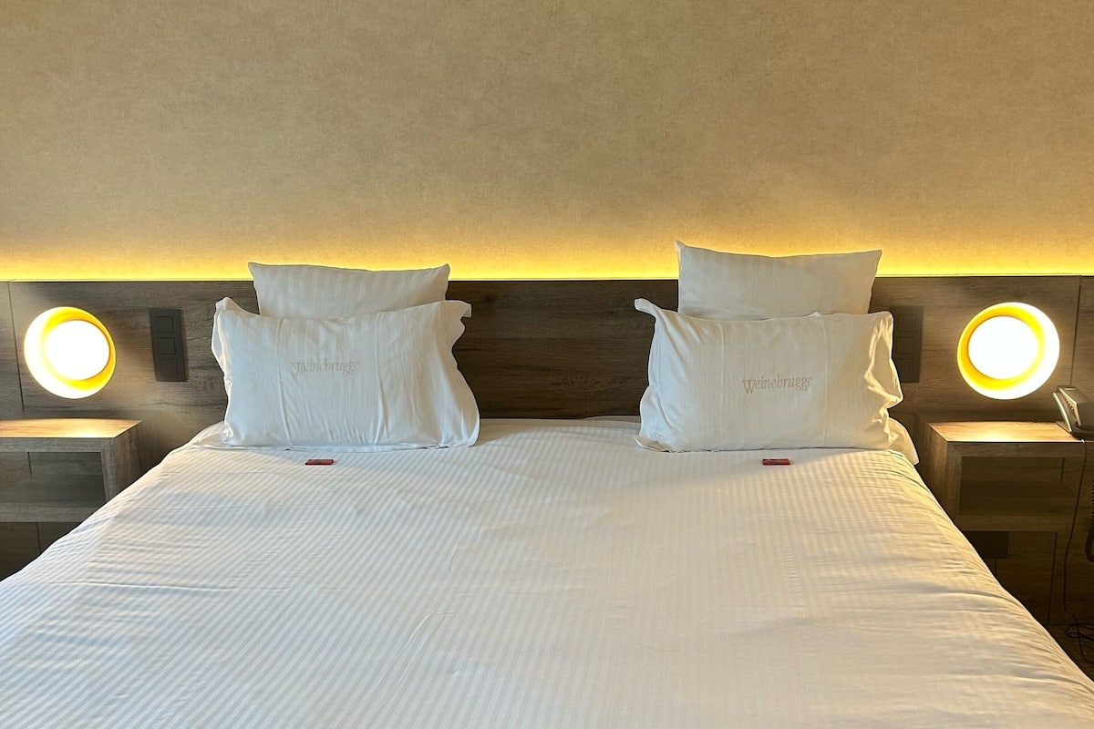 Una cama bien tendida con sábanas y almohadas blancas, dos mesitas de noche con lámparas iluminadas y un cabecero de madera.