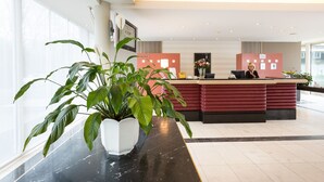 Zona de recepción de hotel con mostrador de mármol, una planta grande en maceta y un recepcionista detrás de un escritorio rojo.