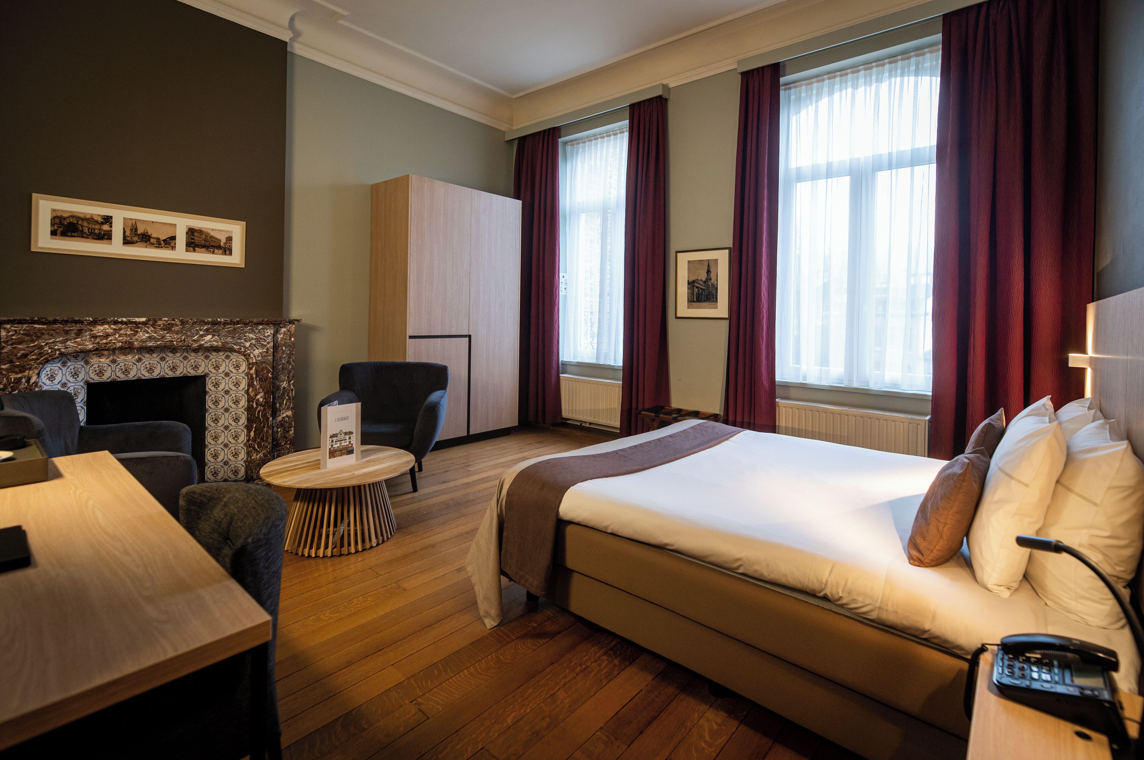 Habitación de hotel con cama, escritorio, silla, mesita redonda pequeña, chimenea y ventanas grandes con cortinas.
