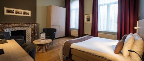 Habitación de hotel con cama, escritorio, silla, mesita redonda pequeña, chimenea y ventanas grandes con cortinas.