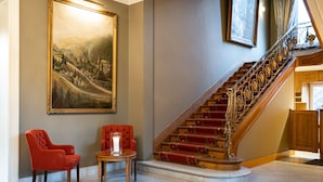 A grand staircase with red carpeted steps and ornate metal railings, flanked by two red armchairs and a small table with a candle.