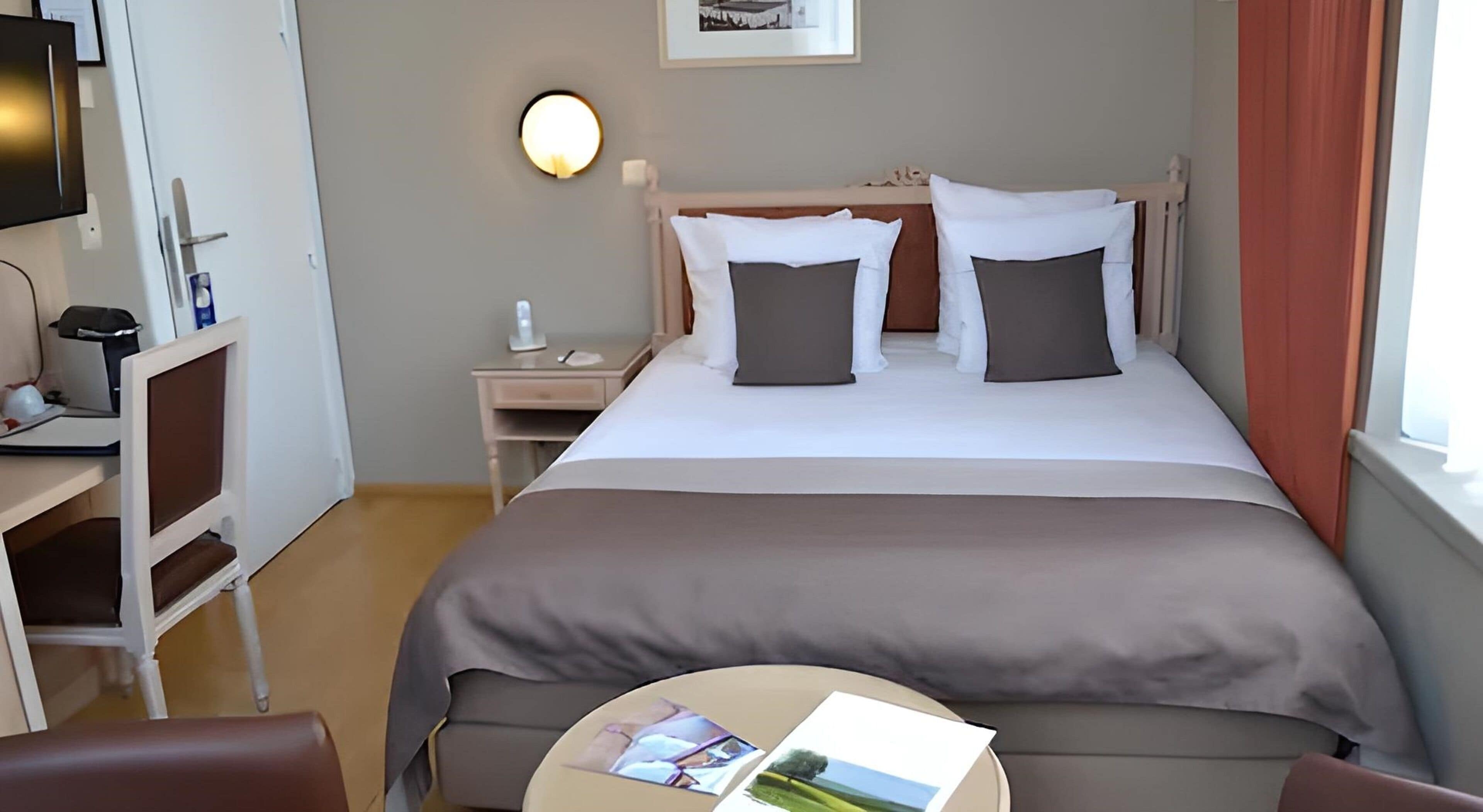 A hotel room with a bed, bedside table, lamp, chair, and a small table with magazines.