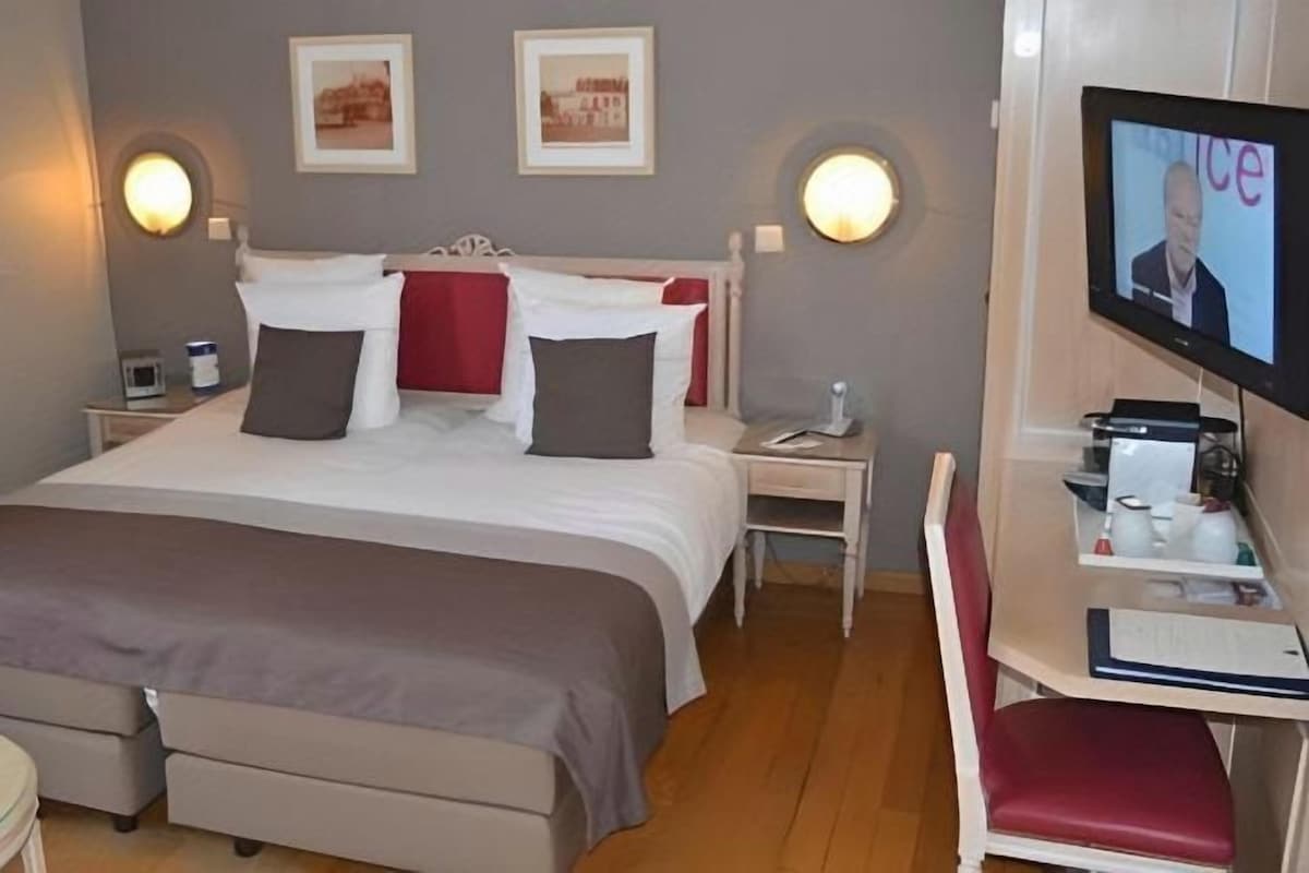 A hotel room with a bed, bedside tables, a desk with a chair, a television, and a small table with a lamp.