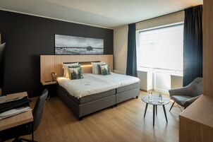 Una habitación de hotel moderna con una cama grande, mesitas de noche, un escritorio, una silla y un ventanal con cortinas.