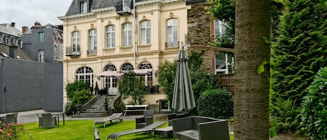 A courtyard with outdoor seating, a stone building with a balcony, and a tree.