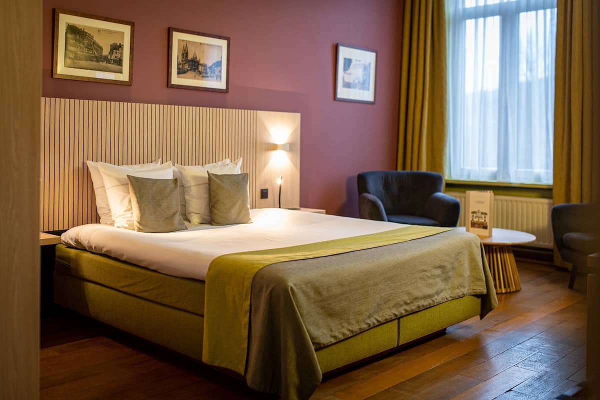 A hotel room with a bed, a chair, a small table, and a window with curtains.