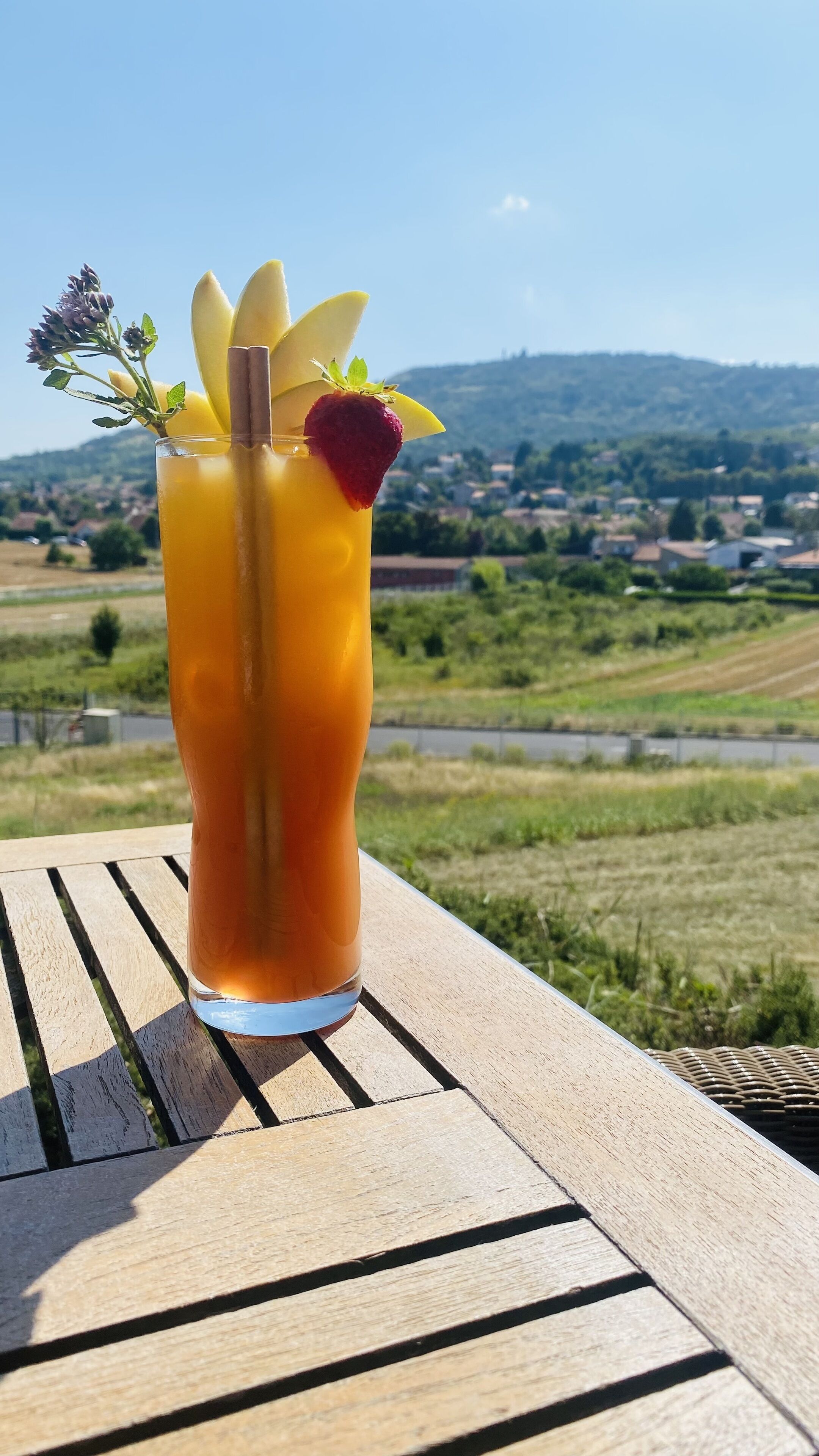 A tall glass of orange beverage with a strawberry and mint garnish on a wooden table with a scenic view of a rural landscape in the background.