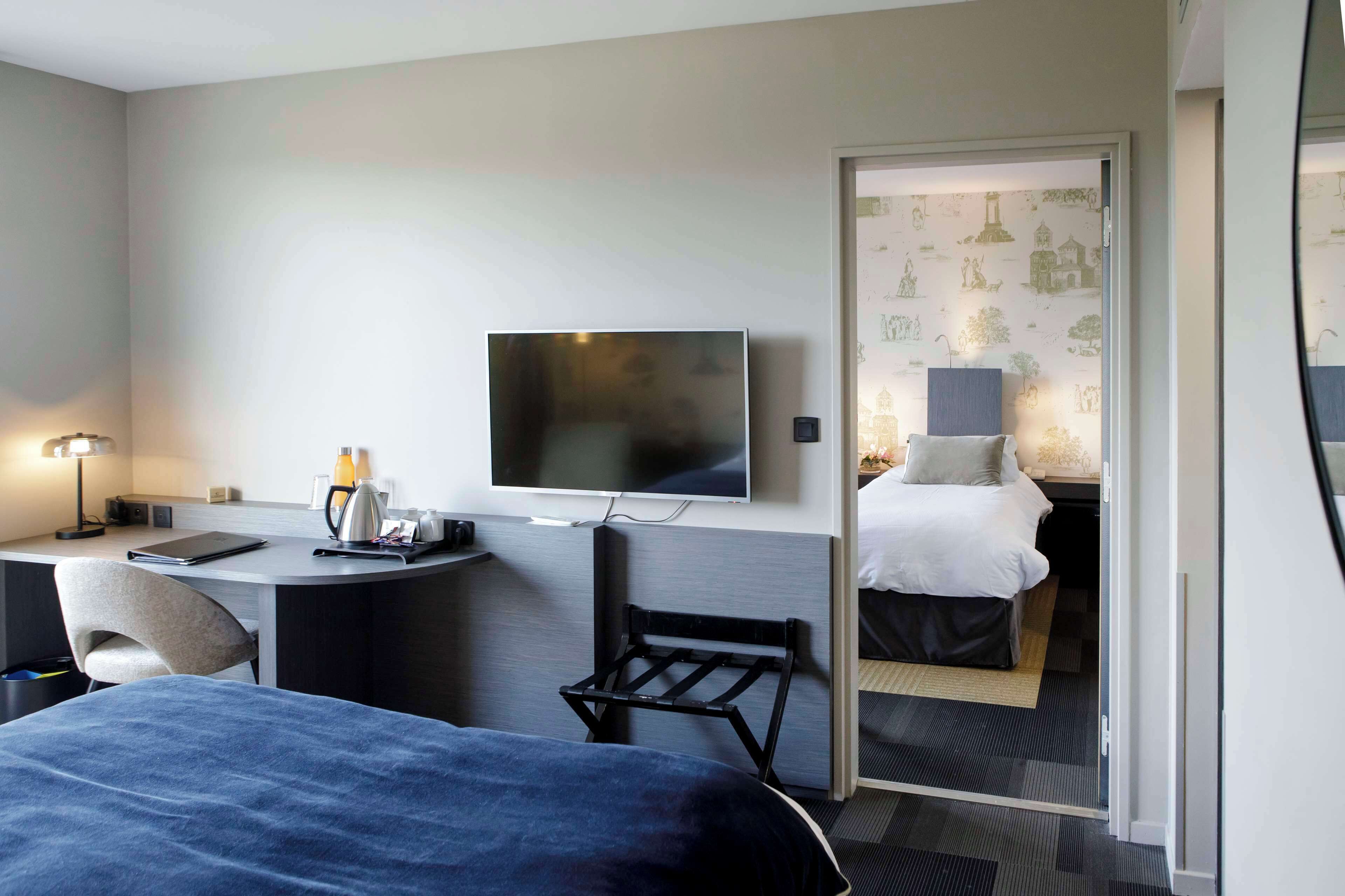 A hotel room with a bed, desk, chair, TV, and a separate bedroom with a bed and nightstand.