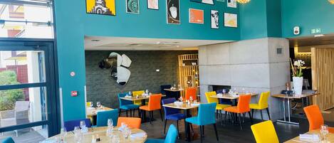 A restaurant with colorful chairs and tables, a wall with artwork, and a view of the outside.