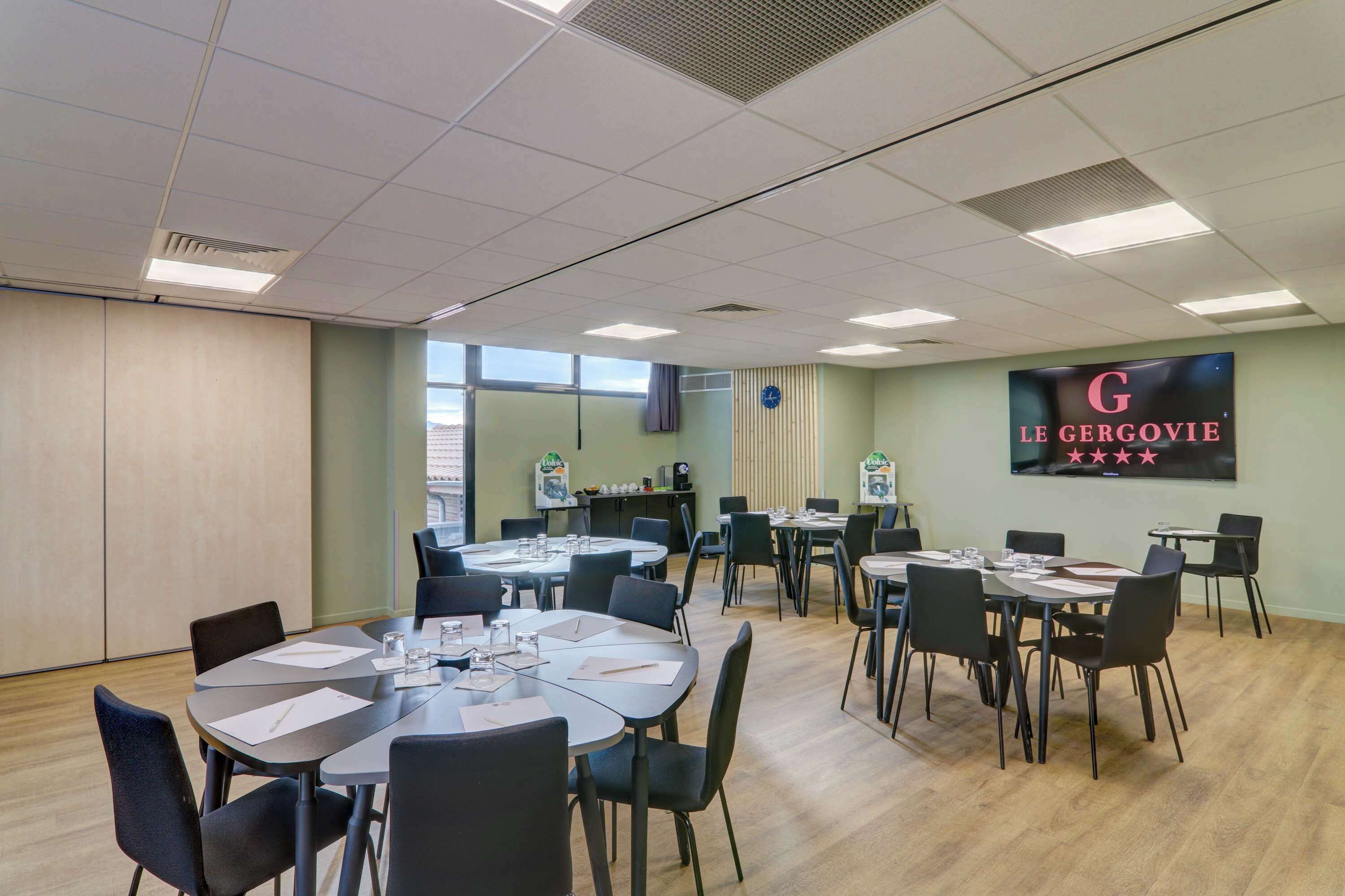 A conference room with multiple tables and chairs, a large screen displaying 'LE GERVIE' with a five-star rating, and a window providing natural light.