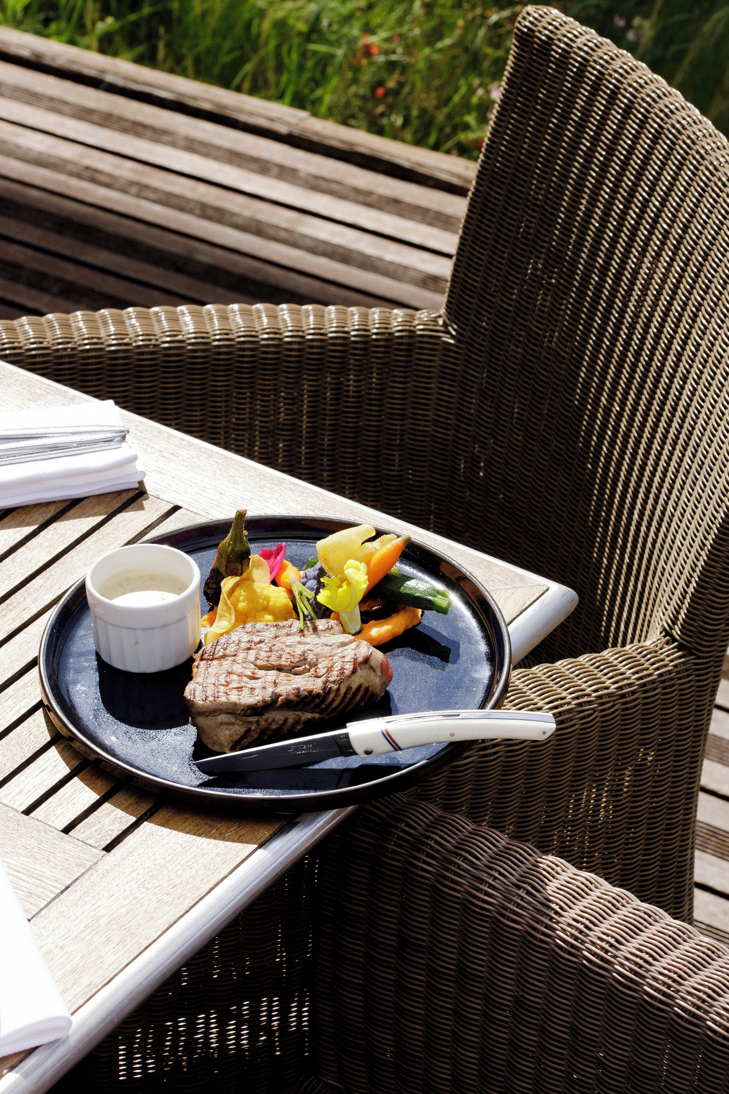 Una silla de mimbre con un plato de comida, que incluye carne y verduras, sobre una mesa de madera.