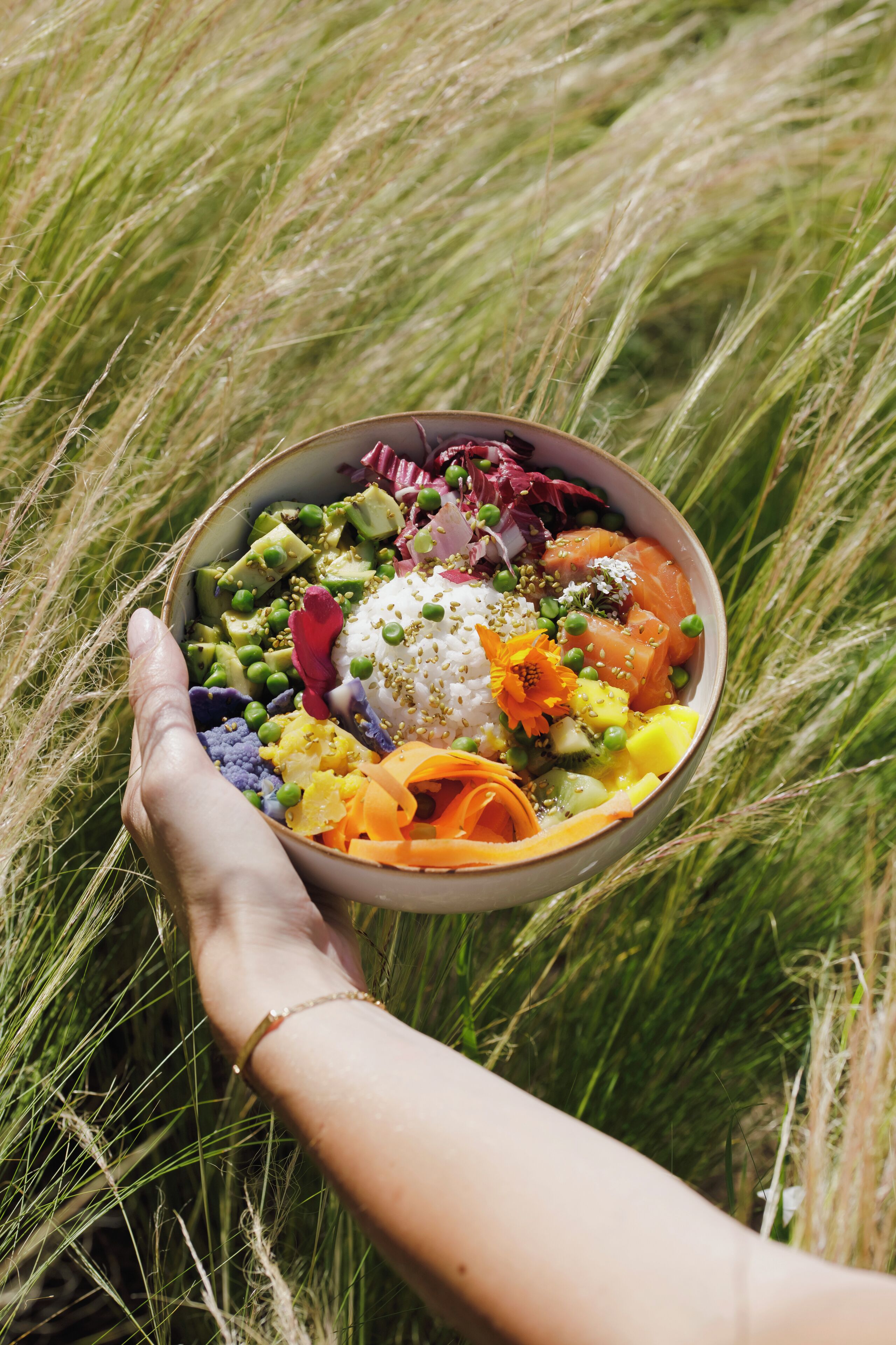 A bowl of colorful food held by a hand amidst tall grass.