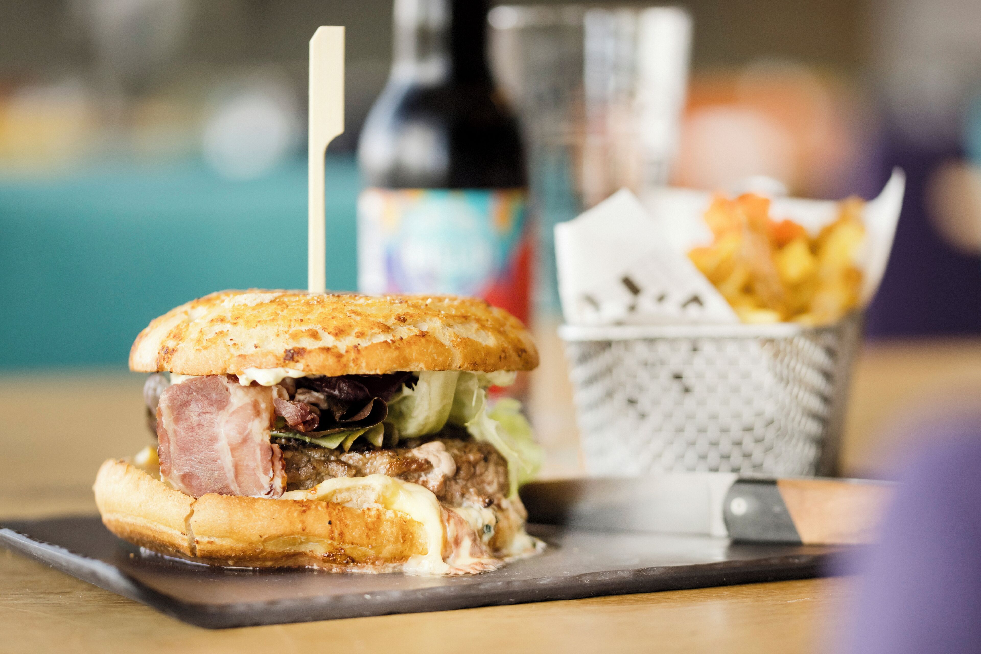 A burger with a bun, lettuce, and a patty, accompanied by a basket of fries and a bottle of sauce.