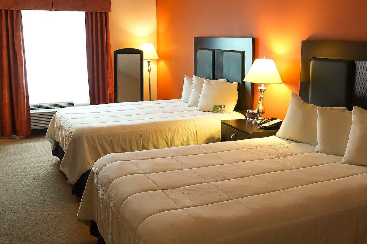A hotel room with two beds, a nightstand with a lamp, a mirror, and a window with curtains.