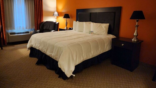 A hotel room with a large bed, two bedside tables, a chair, a window with curtains, and an air conditioning unit. - Grand Hotel  (Spring City)