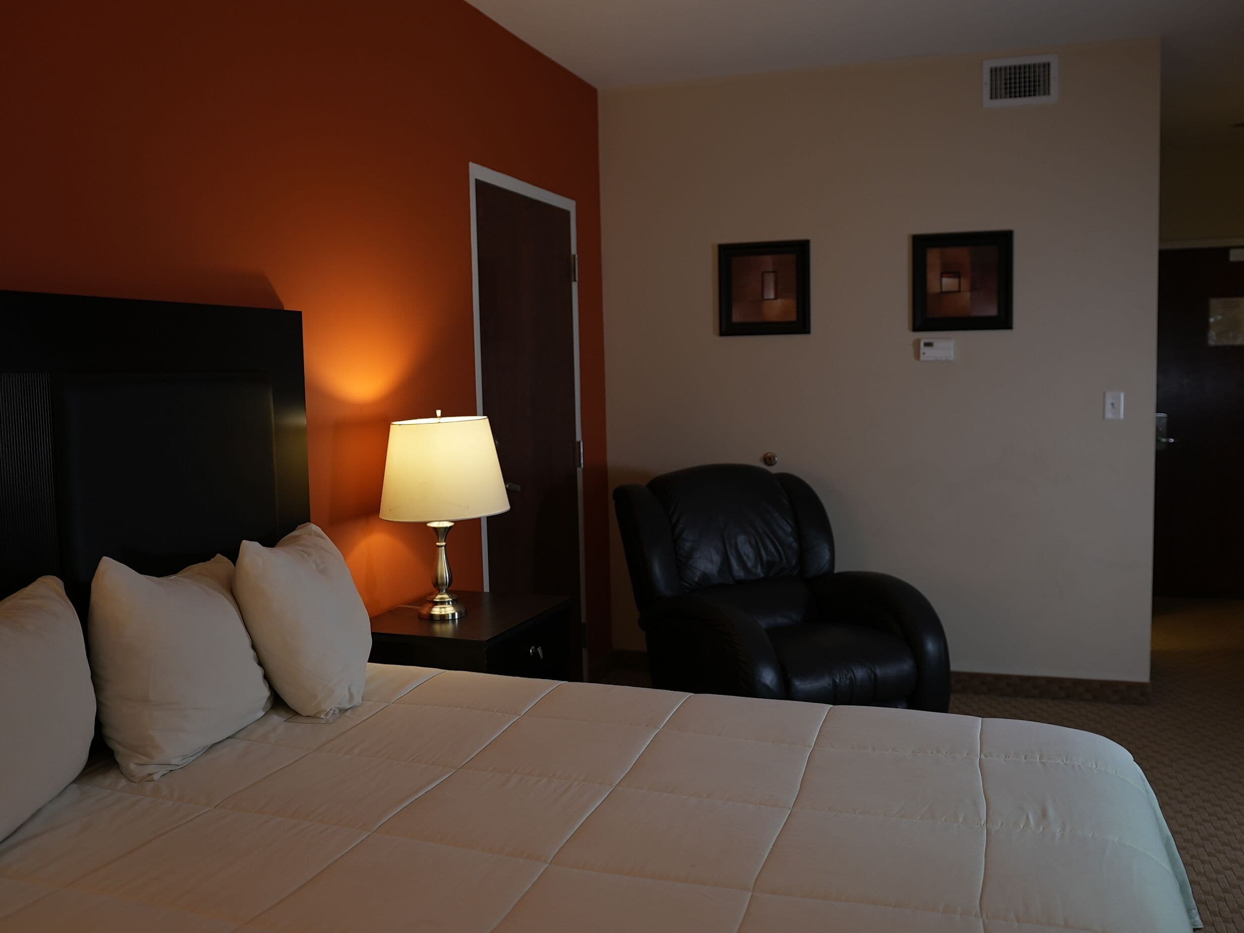 A hotel room with a bed, a nightstand with a lamp, a black leather chair, and two framed pictures on the wall.