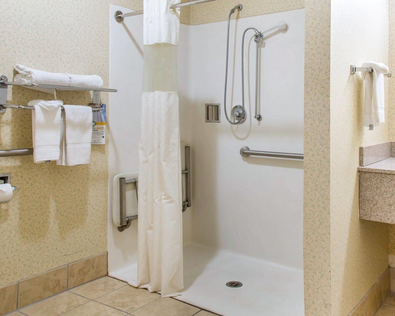 A handicap-accessible shower stall with grab bars, a white shower curtain, and a towel rack with folded towels.