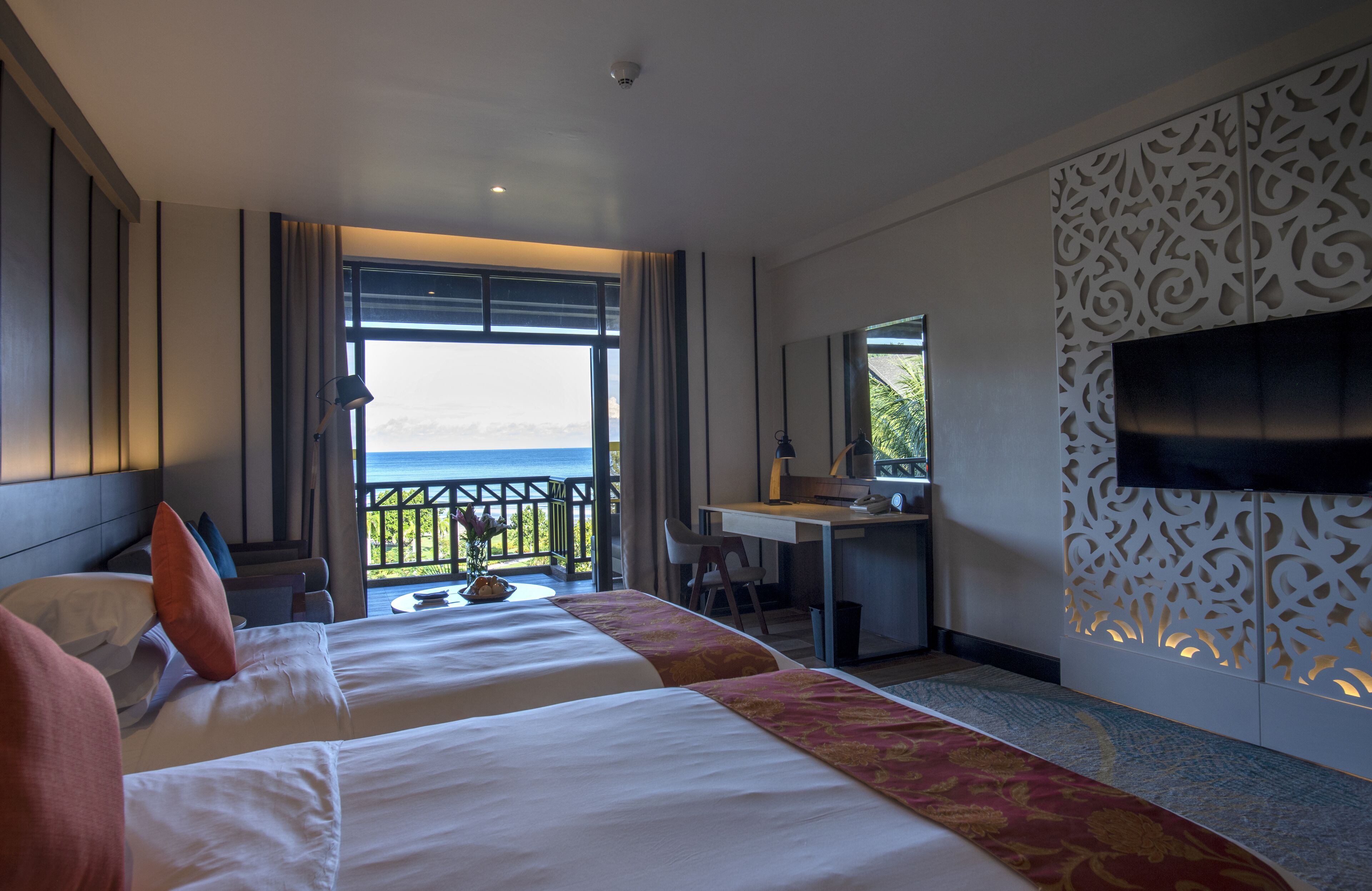 A hotel room with two beds, a desk, a TV, and a balcony with a view of the sea.