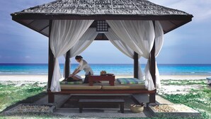 Un kiosque au bord de la plage, doté d’un toit de chaume, de rideaux blancs et d’une table de massage avec des coussins.