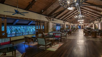 A spacious lounge with wooden floors, comfortable seating, and large windows overlooking palm trees.