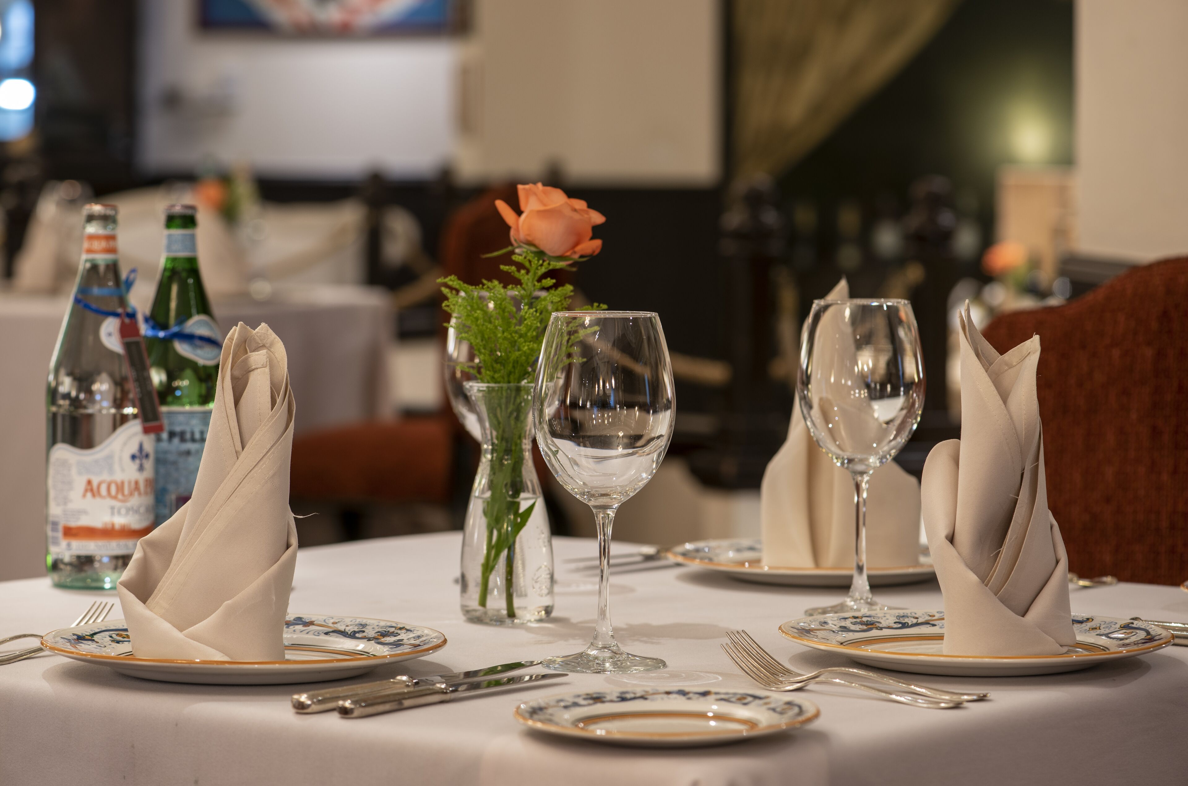 Una mesa de comedor dispuesta con platos, cubiertos y servilletas dobladas. Hay dos botellas de agua y un florero con una rosa.