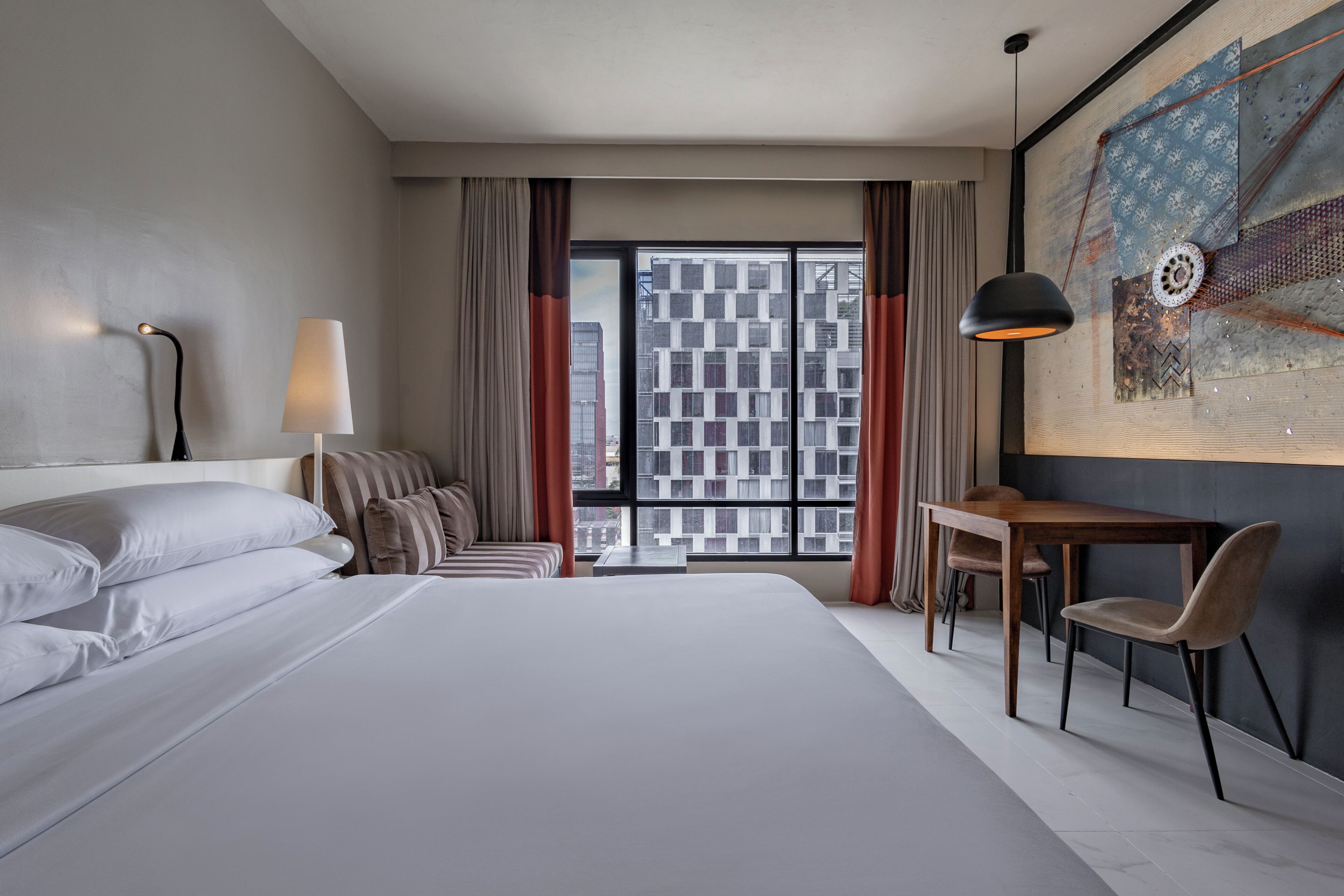 A hotel room with a large bed, a desk, a chair, a lamp, and a view of buildings through the window.